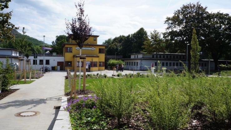 dergrund rechts eine blühende Wiese, auf der linken Seite ist ein Weg zu sehen, der zu einem Platz im Hintergrund vor einem gelben Gebäude führt