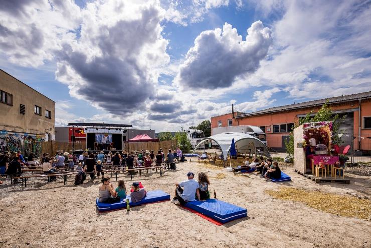 Abbildung einer früheren Veranstaltung im Kulturschlachthof Jena, zu sehen sind eine nachgebildete Strandlandschaft mit Menschen auf Luftmatratzen und Sitzbänken, die Sonne scheint durch den leicht bewölkten Himmel, rechts ist ein Zelt aufgespannt und im Hintergrund ist eine Bühne aufgebaut