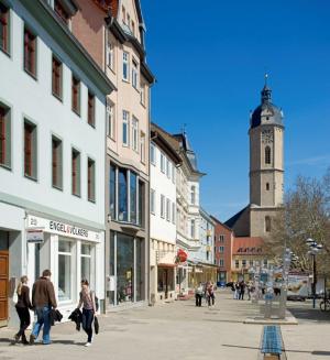 Zu sehen ist die Johannisstraße in Jena. Es ist eine autofreie Einkaufsstraße im Jenaer Zentrum. Auf der linken Seite stehen Mehrfamilienhäuser mit geschäften im Erdgeschoß. Fußgänger schlendern durch die Straße. Am Ende der Straße ist die Stadtkirche mit ihrem hohen Turm zu sehen. 
