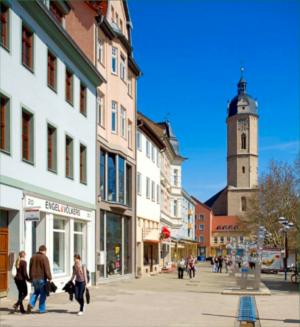 Zu sehen ist die Johannisstraße- eine Fußgängerzone und Einkaufsstraße in Jena. Auf der linken Seite befinden sich Mehrfamilienhäuser mit Geschäften im Erdgeschoß. Menschen gehen die Straße entlang. Am Ende der Straße ist die Stadtkirche mit Turm zu sehen. er
