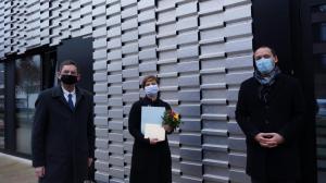 Drei Menschen vor einem Haus mit einer Fassade aus hellgrauem Metall bei der Übergabe einer Urkunde und Plakette für den Fassadenpreis 2020