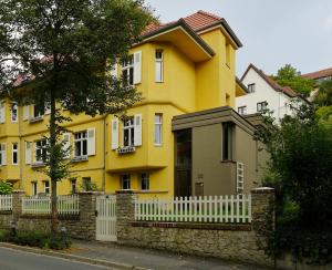 Haus mit gelber Fassade und dunkelgrauem Anbau vor blauem Himmel, im Vordergrund ein Baum und der Gehweg