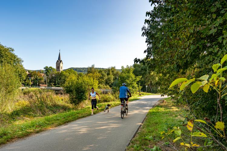 Blick auf einen Radweg mit einem Radfahrer sowie einer Joggerin mit Hund, im Hintergrund ist der Kirchturm von Kunitz zu sehen
