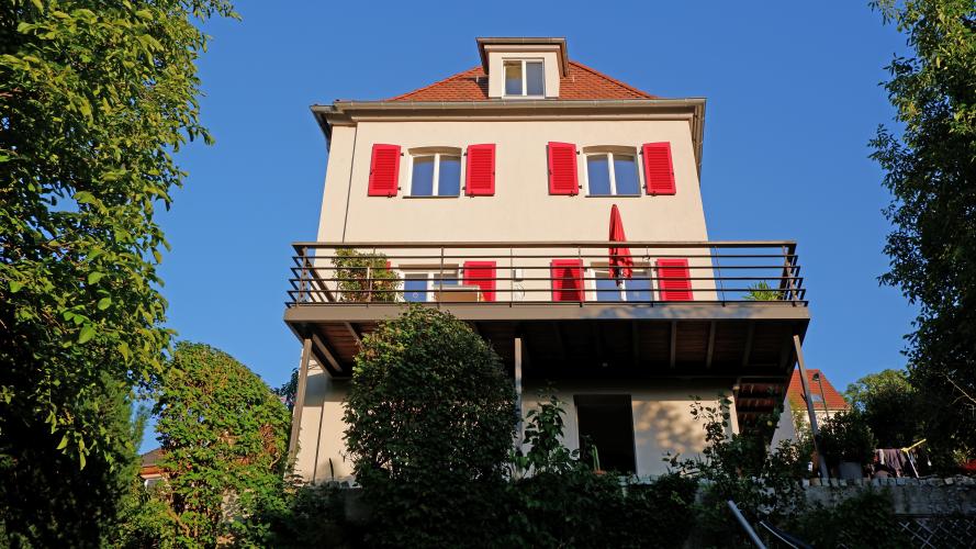 Einfamilienhaus mit heller Fassade, roten Fensterläden aus Holz, großem braunem Balkon und rotem Dach vor blauem Himmel, im Vordergrund Bäume, Büsche und Blumen