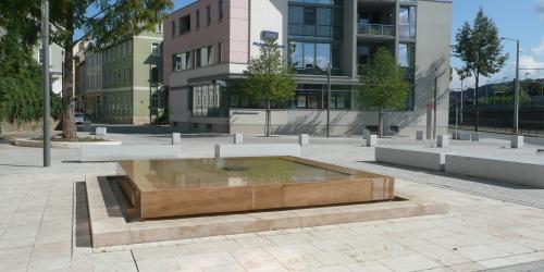 Rechteckiger Brunnen auf einer Freifläche, im Hintergrund ein Gebäude und Bäume 