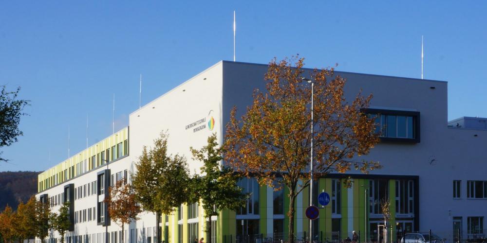 Blick auf die Gemeinschaftsschule Wenigenjena mit grün-weißer Fassade, im Vordergrund eine Baumreihe
