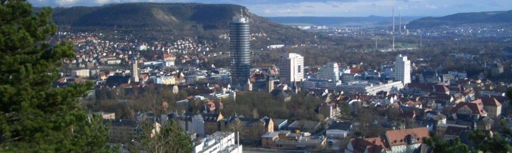 Blick auf die Stadt Jena. Zu sehen sind viel Gebäude und in der Mitte der Intershop-Tower, der höchste Turm in der Innenstadt. Die Stadt wird von Bergen umgeben und ein blauer Himmel mit weißen Wolken ist über der Stadt.