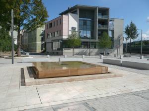 Rechteckiger Brunnen auf einer Freifläche, im Hintergrund ein Gebäude und Bäume 