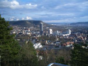 Blick auf die Stadt Jena. Zu sehen sind viel Gebäude und in der Mitte der Intershop-Tower, der höchste Turm in der Innenstadt. Die Stadt wird von Bergen umgeben und ein blauer Himmel mit weißen Wolken ist über der Stadt.