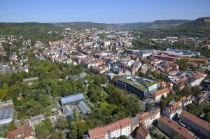 Blick auf Jena. Zu sehen sind eine Vielzahl von Gebäuden - Wohnhäuser und Industriegebäude. Am Horizont ist ein Streifen blauer Himmel zu sehen. Auf der linken Bildseite ist viel Grün in Form von Bäumen zu sehen. Auf beiden Seiten und im Hintergrund wird die Stadt von Bergen eingerahmt.