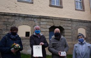 Vier Menschen vor einem Haus mit gelb-grauer Fassade bei der Übergabe einer Urkunde und Palette als Anerkennung beim Fassadenpreis