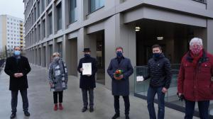 Sechs Menschen vor einem Haus mit heller Fassade und großen Fenstern bei der Übhergabe einer Urkunde und Plakette als Würdigung beim Fassadenpreis