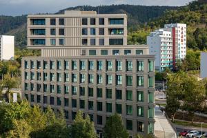 Blick auf ein Hochhaus mit heller Fassade, großen Fenstern und einem Turm, im Hintergrund bewaldete Berge