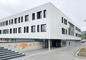 Weißes Haus mit Flachdach, dunklen Fenstern, orangefarbenen Zierelementen und großer Treppe vor bewölktem Himmel, im Vordergrund eine Straße