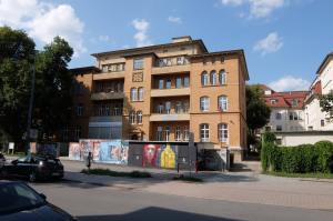 Fotografie des Gebäudes der ehemaligen Frauenklinik mit einer Straße im Vordergrund, einigen parkenden Autos, einer Fußgängerin sowie einer mit Grafittis versehenen Mauer