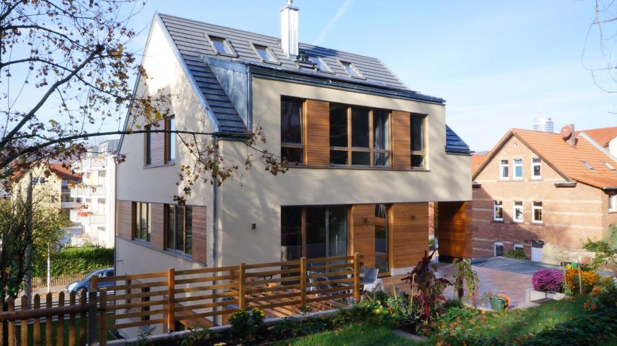 Haus mit weißer Fassade und Holzelementen vor blauem Himmel, im Vordergrund ein Holzzaun und ein Baum