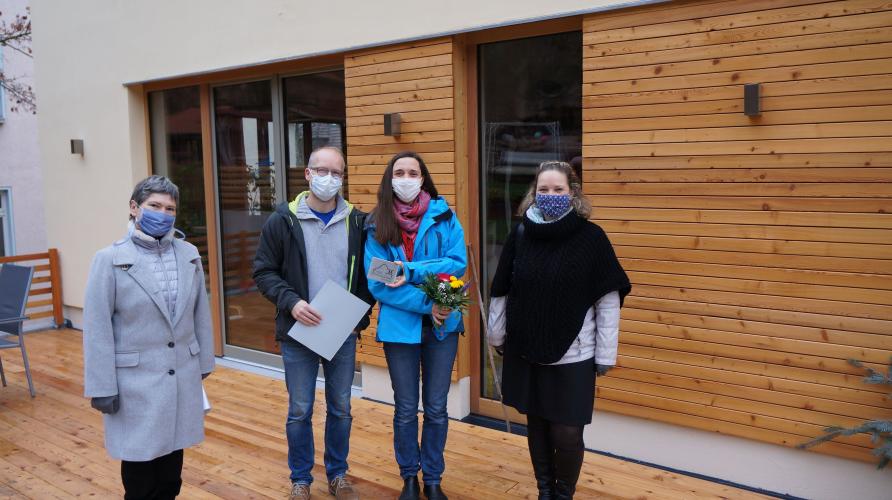 Vier Menschen vor einem Haus mit braun-weißer Fassade bei der Übergabe einer Urkunde und Plakette als Anerkennung beim Fassadenpreis