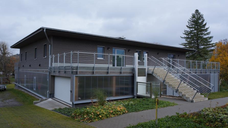 graues Haus mit Flachdach und großer Treppe vor bewölktem Himmel, im Vordergrund Rasen und blühende Pflanzen