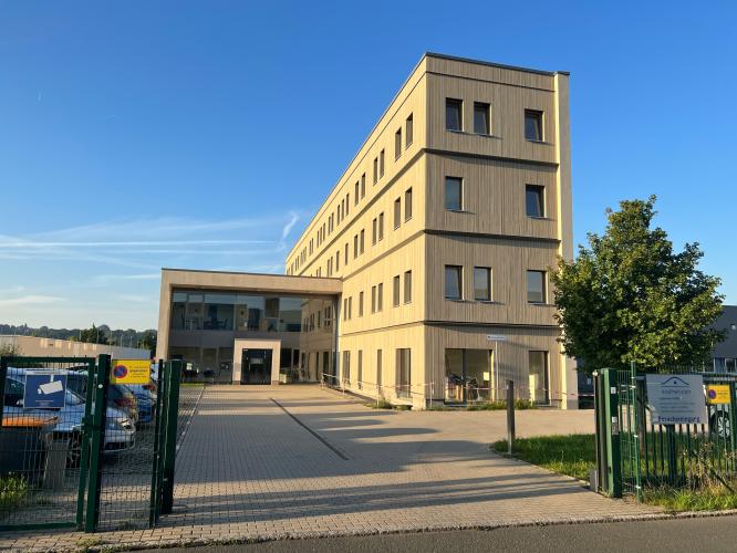 Firmengebäude mit heller Fassade, großen Fenstern, Flachdach und einem kleineren Anbau mit großer Glasfront vor blauem Himmel, davor eine gepflasterte Einfahrt, Autos auf einem Parkplatz, Rasen, ein Zaun und ein Baum