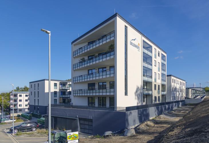 Wohnkomplex aus 4 Gebäuden mit weiß-baleur Fassade und blauem Sockel mit umgebenden Grünflächen vor blauem Himmel