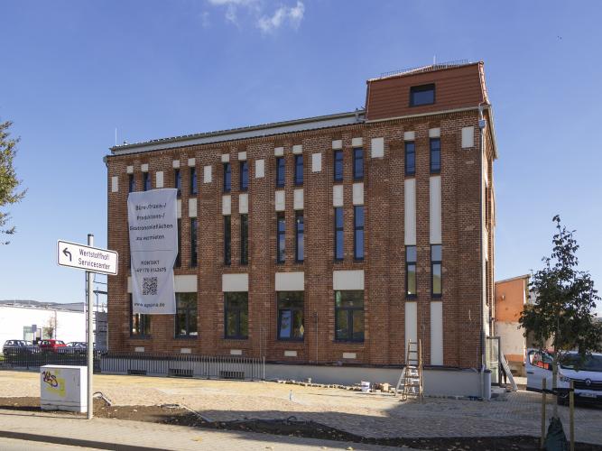 Gewerbebau mit Klinkerfassade, weißen Wandelementen und großen Fenstern, im Vordergrund gepflasterte Stellplätze, dahinter blauer Himmel