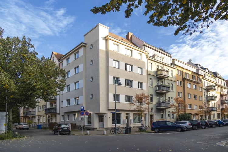 Mehrfamilienhaus mit Fassade in weiß und beige, im Vordergrund Straße mit geparkten Autos und Bäumen, darüber blauer Himmel mit weißen Federwolken