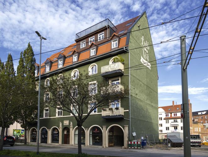 Mehrfamilienhaus mit grüner Fassade, rot-braunem Dach, weißen Fenstern und Torbögen vor blauem Himmel mit weißen Federwolken