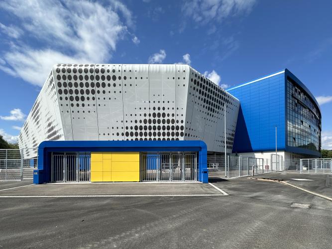 neugebaute Fußballarena mit weiß-blauer Fassade und gelben Elementen am Eingangsbereich mit geteerten Stellplätzen vor blauem Himmel mit Federwolken