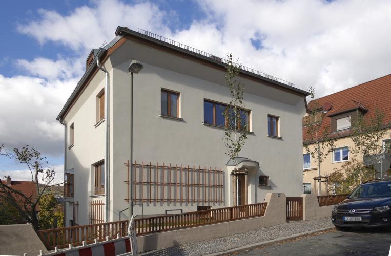 Einfamilienhaus mit weißer Fassade und braunen Holzelementen, im Vordergrund eine Straße und ein niedriger Zaun