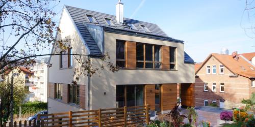 Haus mit weißer Fassade und Holzelementen vor blauem Himmel, im Vordergrund ein Holzzaun und ein Baum