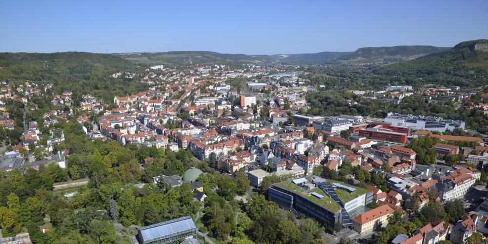 Zu sehen ist ein Blick auf Jena. Im Vordergrund und in der rechten Bildhälfte sind Häuser in dichter Bebauung zu sehen. In der Bildmitte und auf der rechten Seite sind das Planetarium und der Botanische Garten von Bäumen umrahmt zu sehen. Am Horizont ist ein Streifen blauer Himmel zu sehen.