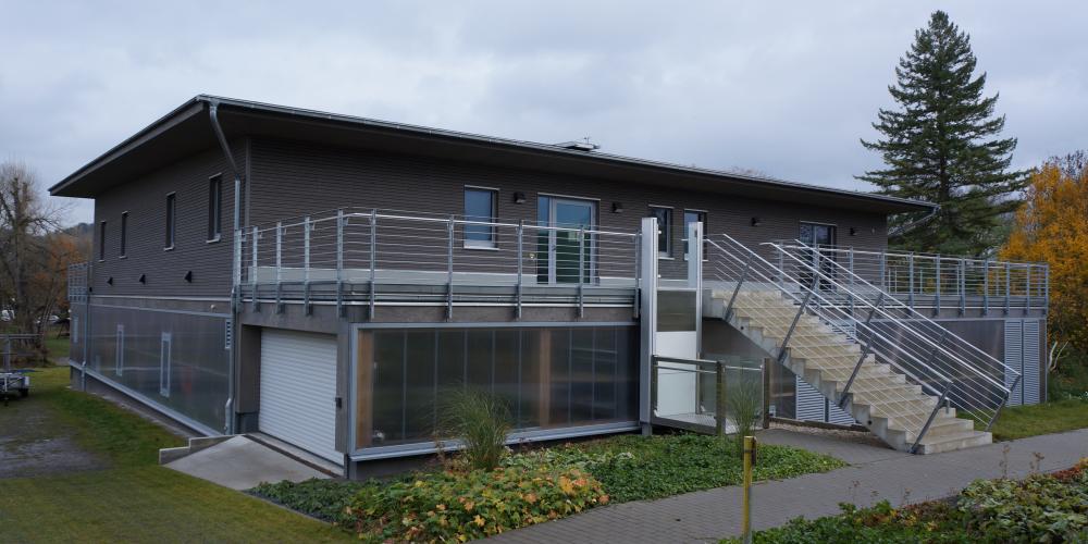 graues Haus mit Flachdach und großer Treppe vor bewölktem Himmel, im Vordergrund Rasen und blühende Pflanzen