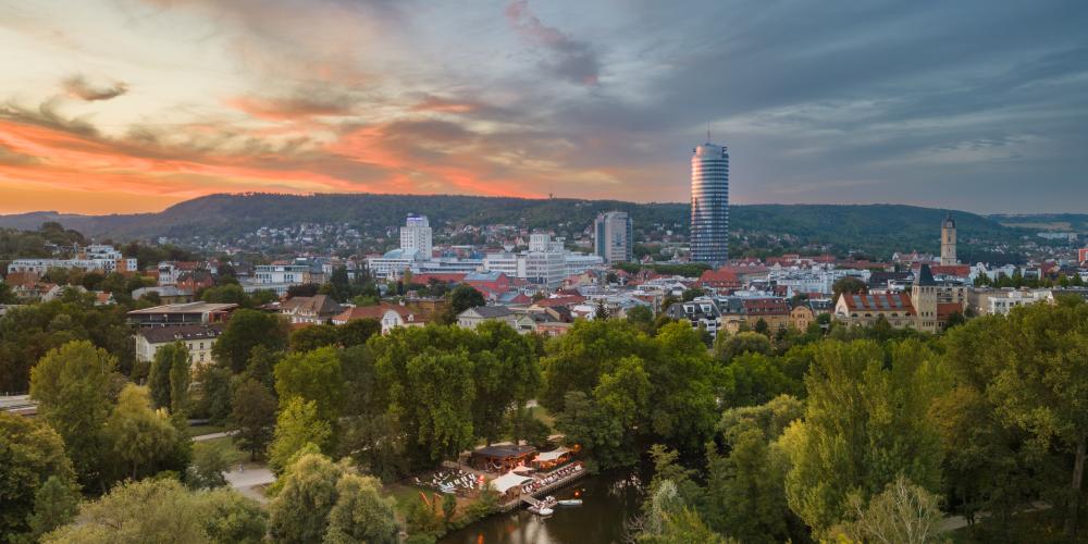 Panoramaaufnahme der Stadt Jena mit einem Fluss und viel Grün runsherum im Vordergrund und Bebauung im Hintergrund