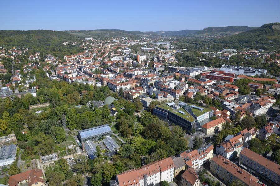 Zu sehen ist ein Blick auf Jena. Im Vordergrund und in der rechten Bildhälfte sind Häuser in dichter Bebauung zu sehen. In der Bildmitte und auf der rechten Seite sind das Planetarium und der Botanische Garten von Bäumen umrahmt zu sehen. Am Horizont ist ein Streifen blauer Himmel zu sehen.