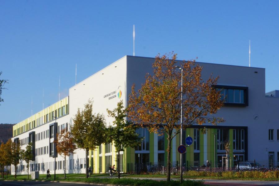 Blick auf die Gemeinschaftsschule Wenigenjena mit grün-weißer Fassade, im Vordergrund eine Baumreihe