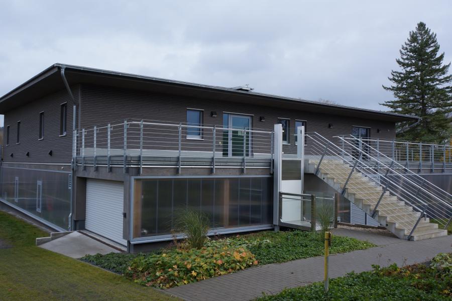 graues Haus mit Flachdach und großer Treppe vor bewölktem Himmel, im Vordergrund Rasen und blühende Pflanzen