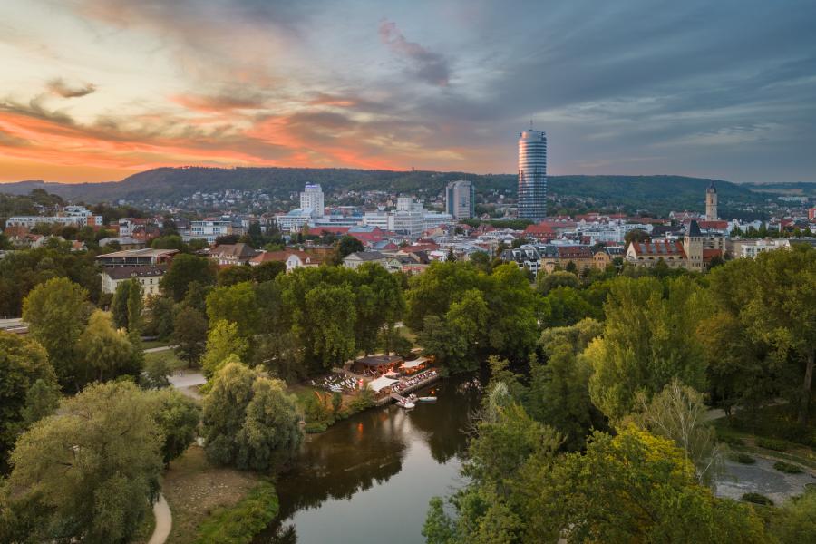 Panoramaaufnahme der Stadt Jena mit einem Fluss und viel Grün runsherum im Vordergrund und Bebauung im Hintergrund